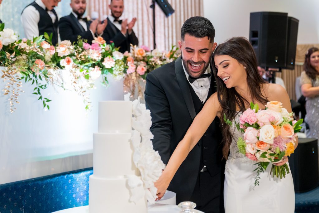 Laughing bride and groom cutting their three-tier white wedding cake in front of a flower-covered head table.